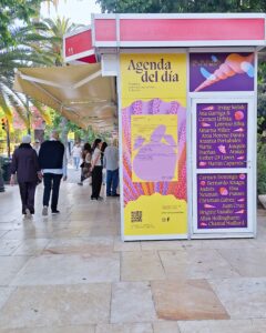 End view of a stall at an outdoor bookview. It's covered in posters listing the author participants. People can be seen strolling and browsing in front of the stalls. Palm trees are on eother side of the stall.