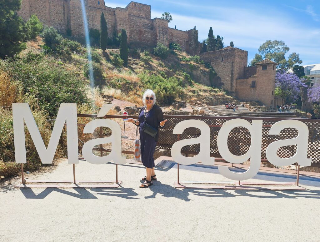 A woman in a blue dress and white hair poses in the sign that says Malaga. Behind her is the ancient stone Arab fortress and ruins of a Roman theater.