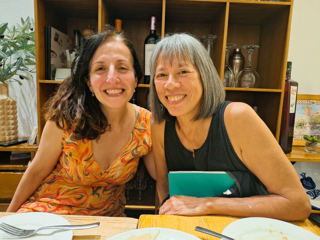 Two smiling women sitting at a table in a restaurant. Behind them is a shelf of wine bottles and glasses. In front of them are empty plates.