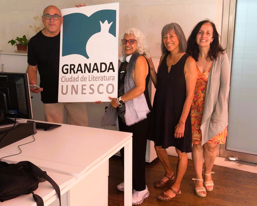 A man with glasses hold up one side of a sign that says Granada Ciudad de Literatura UNESCO. A woman with white hair holds the other side of the sign. Two dark-haired women stans beside her. They are inside an office.