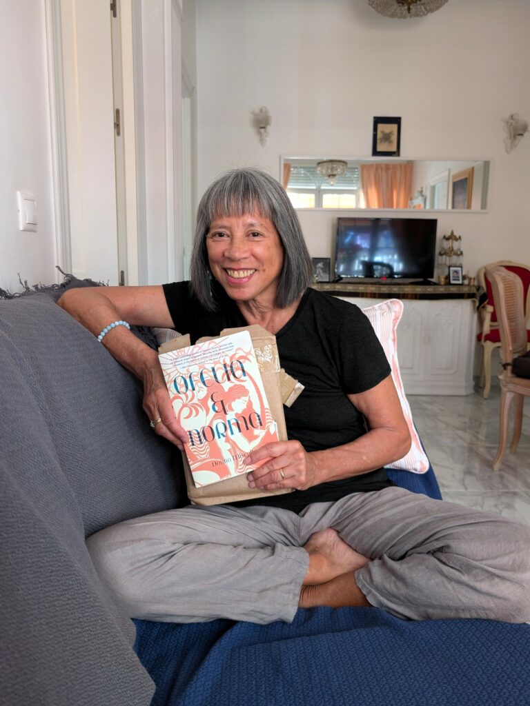 A brown-skinned woman with a pageboy haircut wearing a black T-shirt and gray pants sits cross-legged on a couch holding a book in front of her .