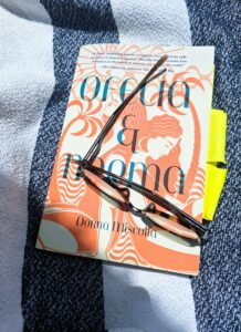 Book titled Ofelia and Norma with a pair of glasses set on top lying on a blue and white striped beach towel.