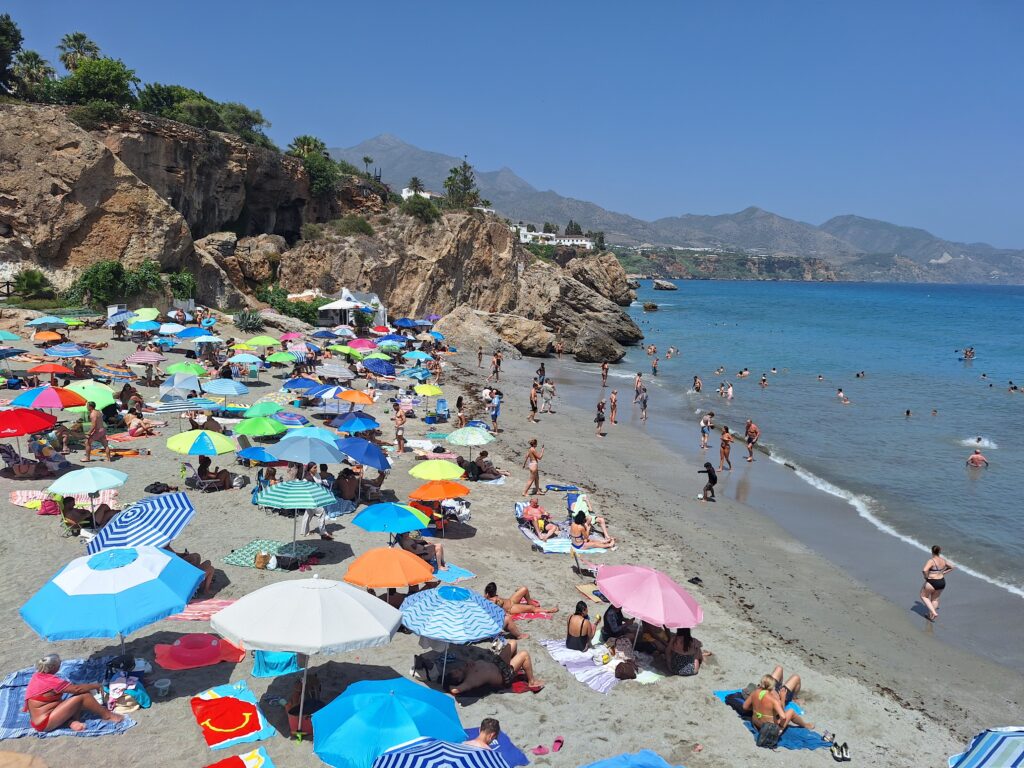 Beach crowded with colorful umbrellas with people in the surf. rocky cliffs separate the beach from other beaches beyond. Town buildings are visible atop the cliff.