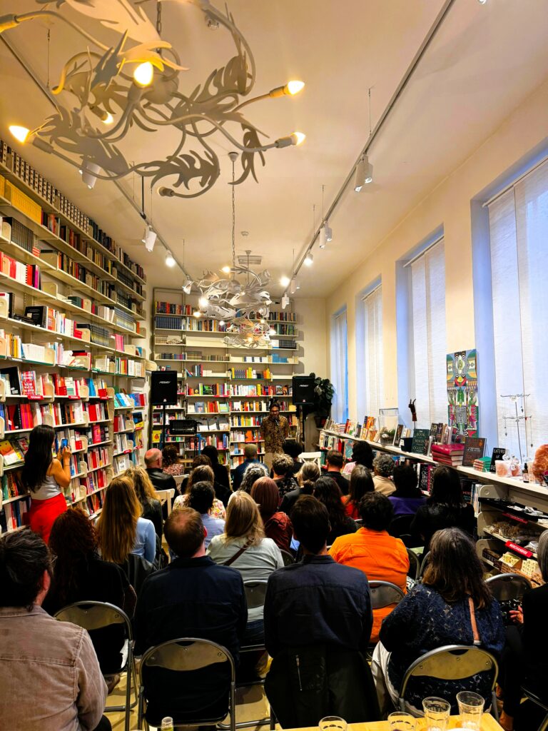 Narrow room full of seated audience listening to a speaker. The wall on the left is lined with books. The wall on the right is a bank of windows.
