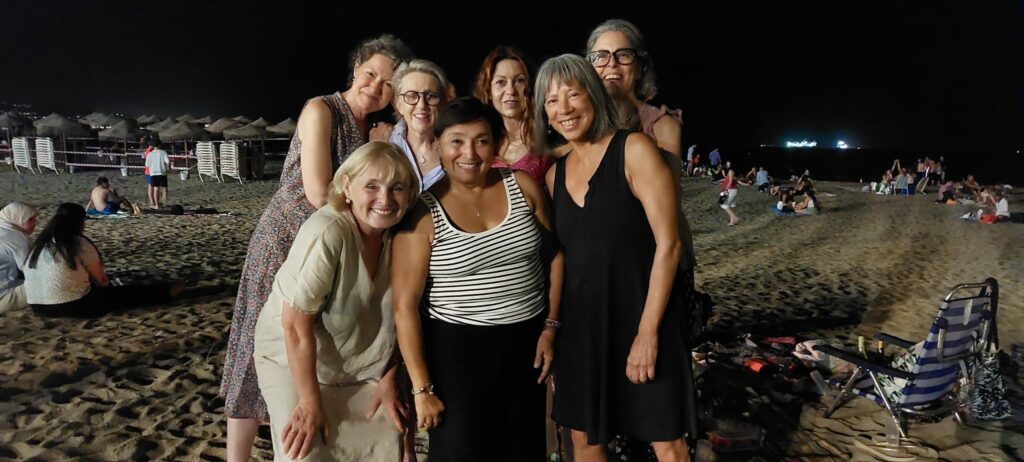 Seven women in two rows pose together on a beach at night.