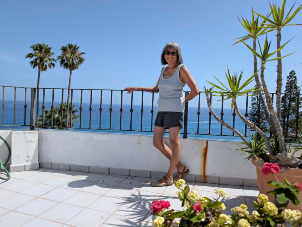 Woman in light gray tank top and darker gray shorts leans against the rail of a garden terrace. Palm trees are visible in the background.