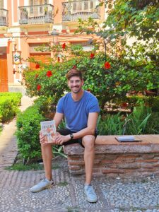 A tall young man with glasses wearing a blue T-shirt and black shorts sits on a stone bench in a courtuard in front of a hibiscus bush in bloom, holding a novel toward the camera.