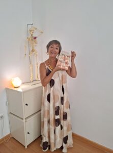 A smiling, dark-haired woman ina long brown and tan spotted sun dress stands next to a skeleton atop a two-drawer credenza. She holds a book up to display its cover.