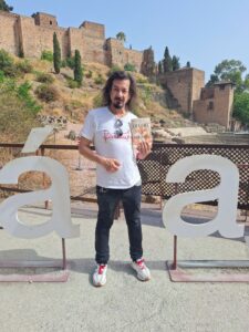 A bearded man with long shoudler-length hair wearing a white T-shirt, dark pants, and white shoes stands with an Arab fortress behind him, holding a book to display its cover.