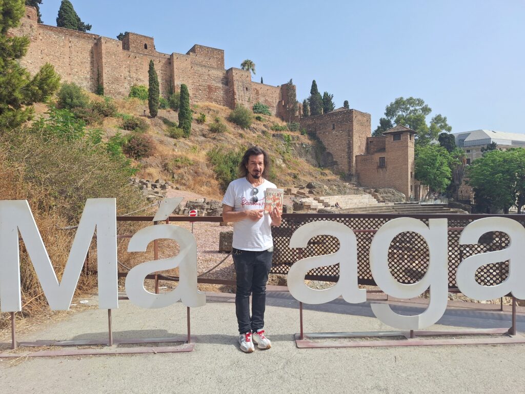 A man in a white shirt and dark pants stands in the middle of the sign that spells Malaga with the Arab fortress in the background as he holds up a book to display its cover.