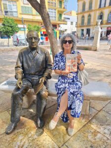 A woman in a blue and white dress and sunglasses sits on a stone bench next to a statue of a sitting Picasso. She holds a book up to display its cover.
