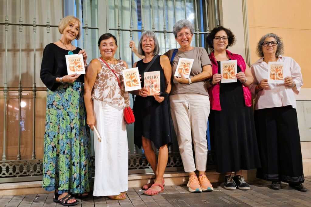 Six women standing and holding a large postcard with a book cover printed on it. The book is Ofelia and Norma by Donna Miscolta